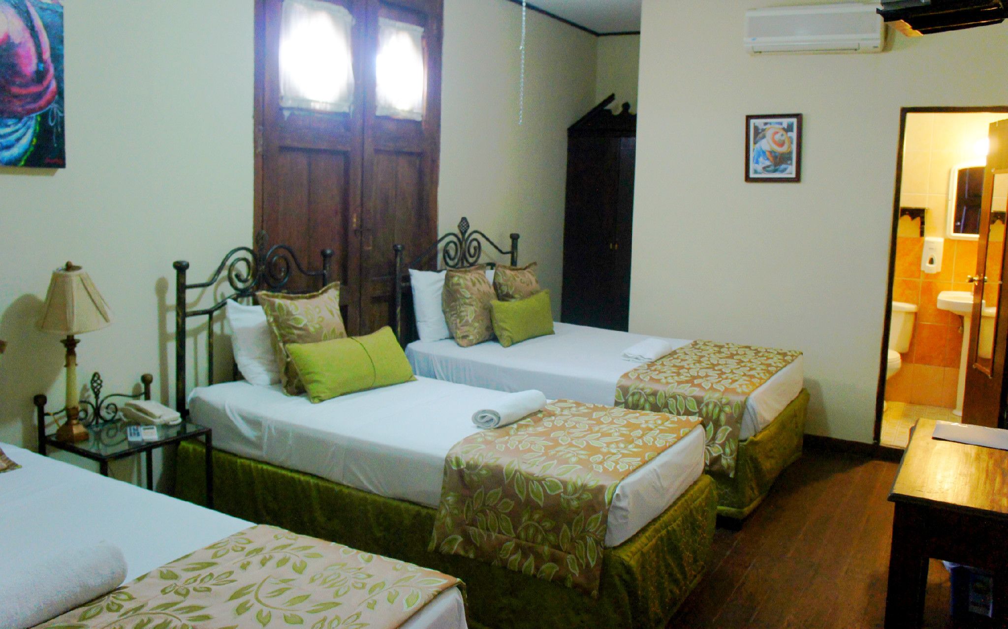 A hotel room with two single beds, a bedside table with a lamp, a wooden wardrobe, and a bathroom visible through an open door.