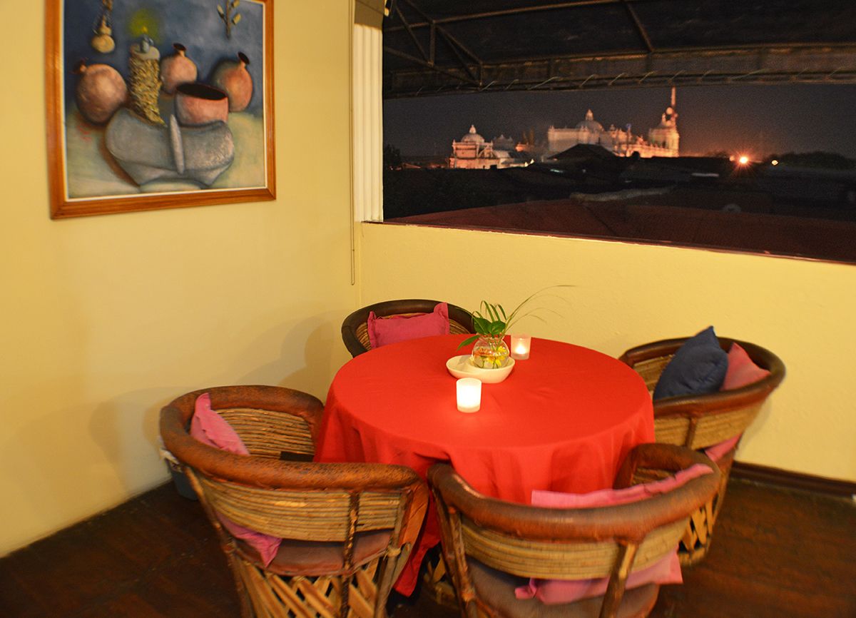 A dining area with wicker chairs and a red tablecloth, a painting on the wall, and a view of a distant building at night.