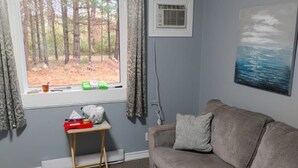 A living room with a grey couch, a small table with items, a window with curtains, an air conditioning unit, and a painting on the wall.