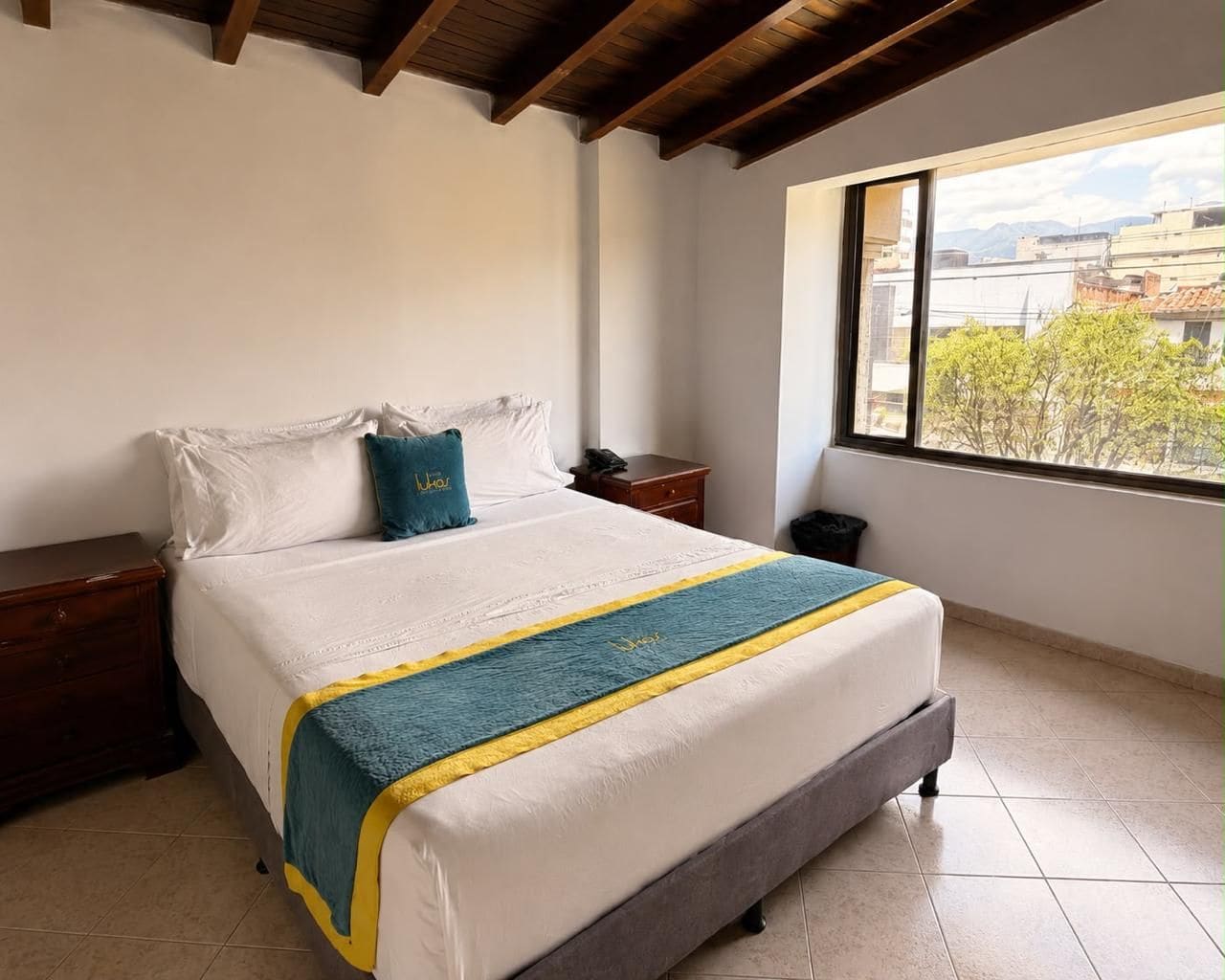 A bedroom with a bed, a nightstand, a bedside table, a window with a view of buildings, and a tiled floor.
