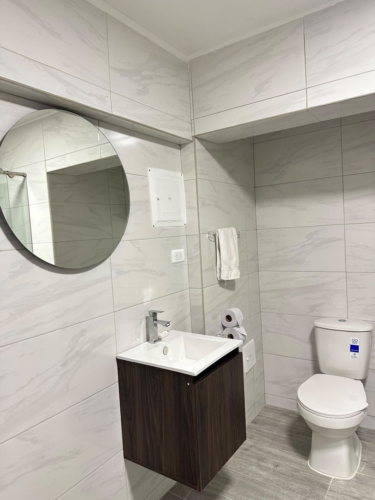 Une salle de bain moderne avec un mur en marbre, un miroir rond, un lavabo blanc, un meuble-lavabo en bois foncé et des toilettes blanches.