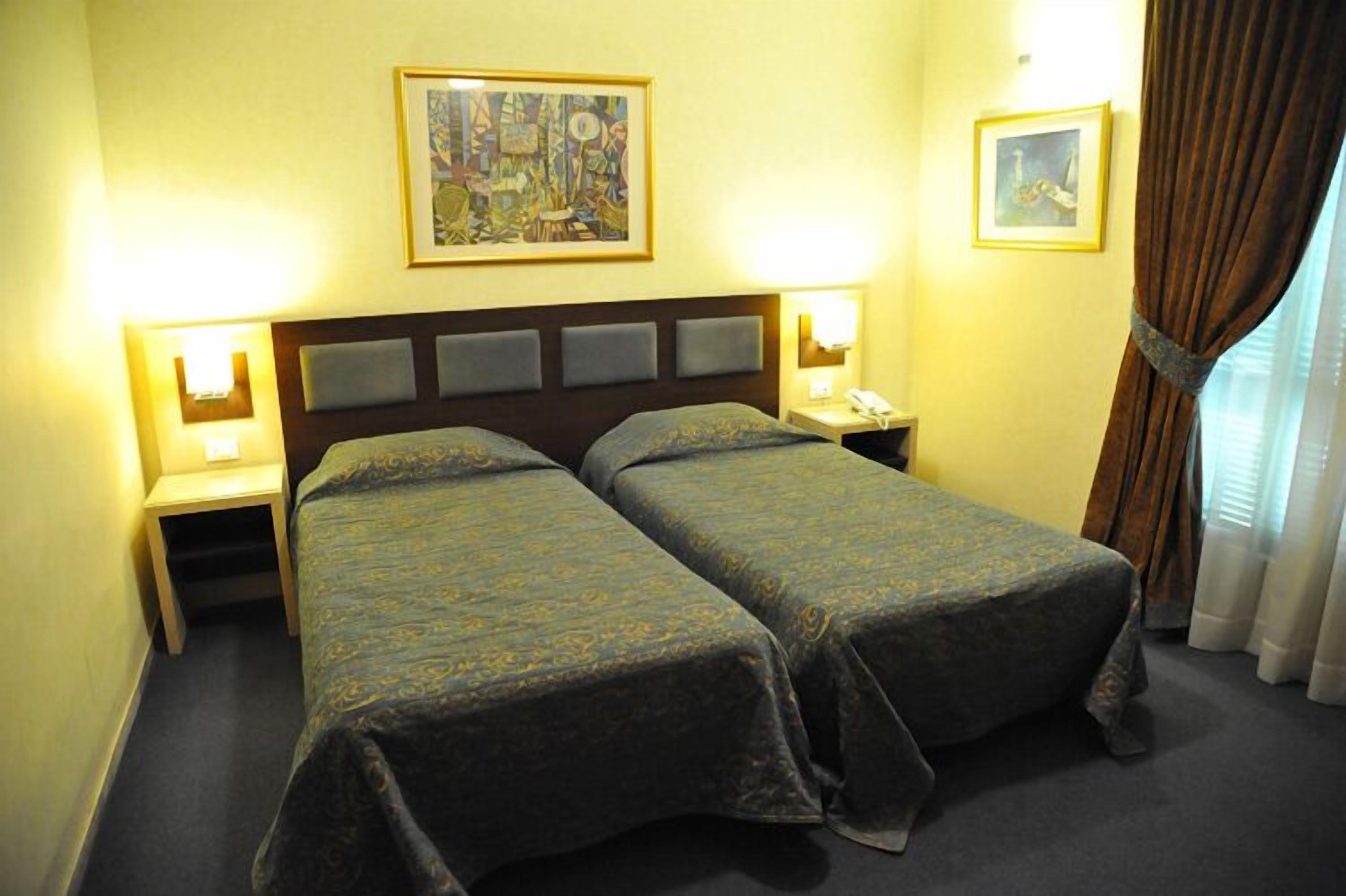 A hotel room with two single beds, a wooden headboard, bedside lamps, and framed artwork on the walls.