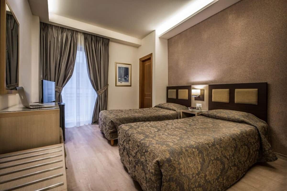 A hotel room with two beds, a desk with a laptop, a TV, and a large window with curtains.