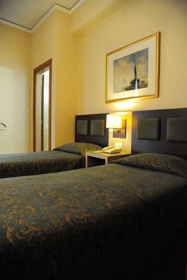 A hotel room with two beds, a nightstand with a lamp, a framed picture on the wall, and a door. - Rex Hotel (Nafplio)