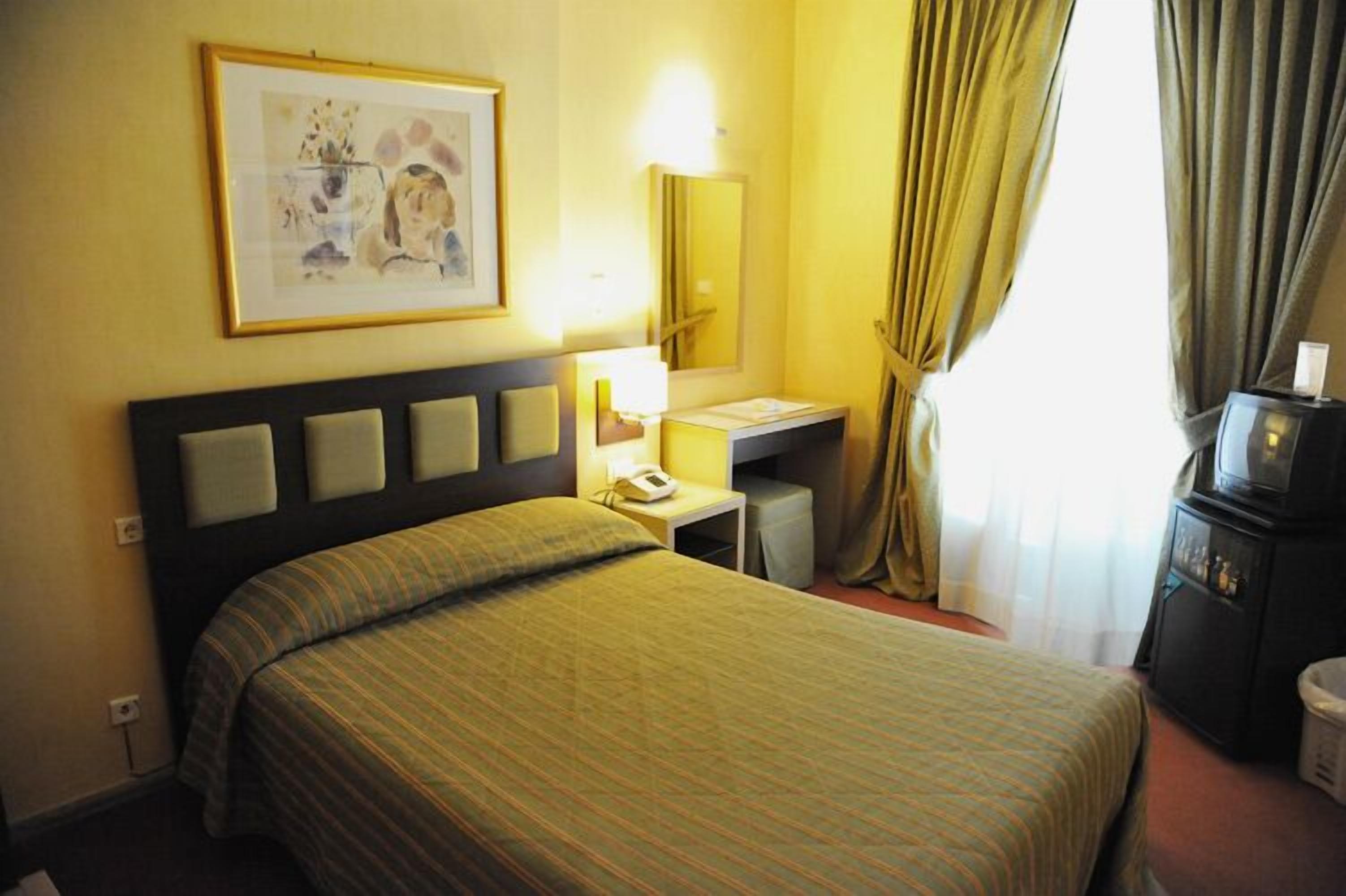 A hotel room with a bed, a bedside table with a lamp, a telephone, a small desk, a mirror, and a television.
