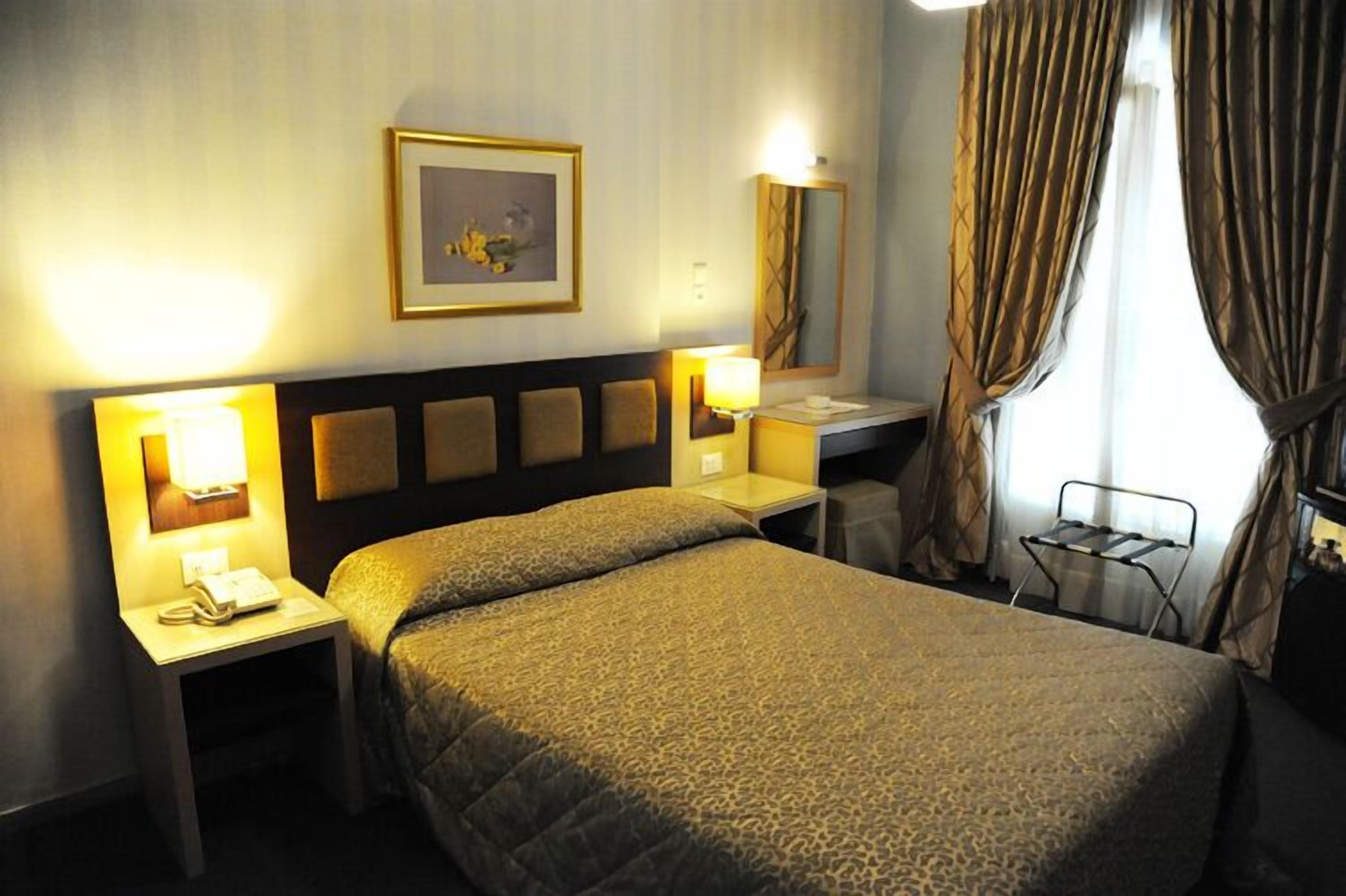 A hotel room with a bed, bedside lamps, a nightstand, a desk, a chair, a mirror, and a window with curtains.