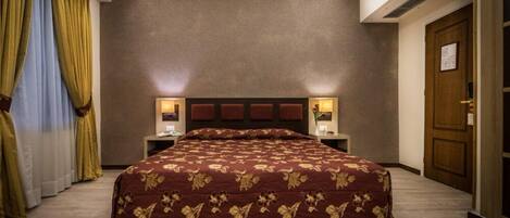 Quarto de hotel com uma cama grande, duas luminárias de cabeceira, um painel de madeira na cabeceira e uma porta à direita.