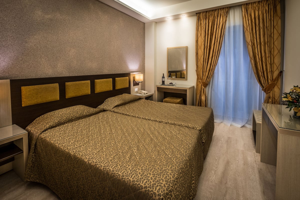 A hotel room with a large bed, a bedside table, a desk, a mirror, and a window with curtains.