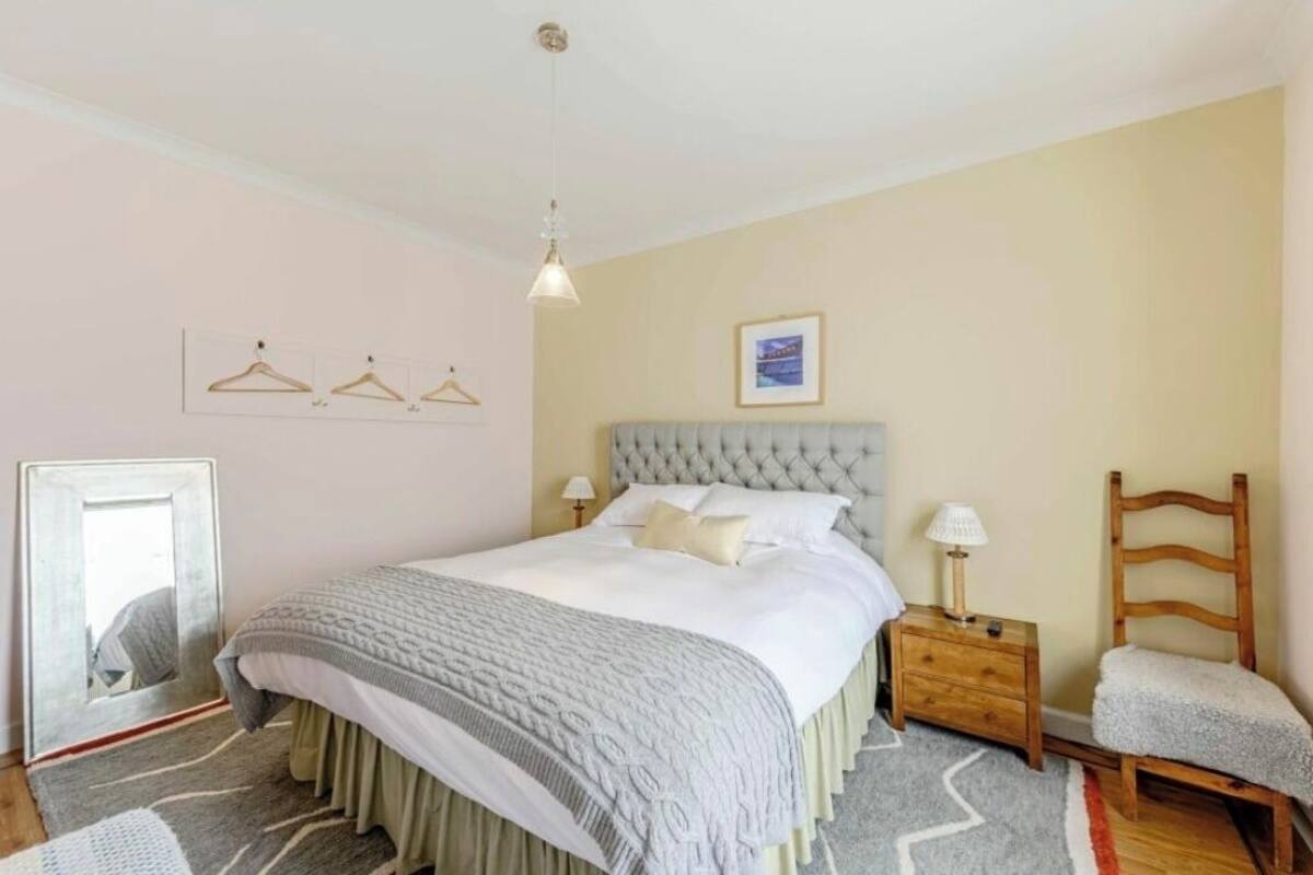 A bedroom with a tufted headboard, a bed with a grey and white patterned bedspread, a wooden nightstand with a lamp, and a wooden chair.