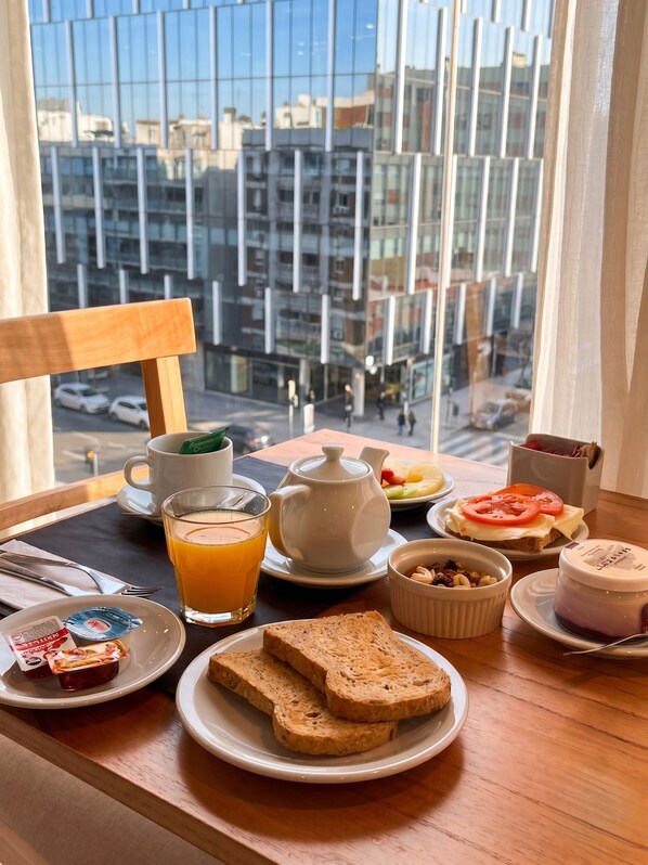 Bandeja de desayuno con tostadas, fruta, té y un pequeño recipiente con frutos secos sobre una mesa junto a una ventana con vistas a la ciudad.