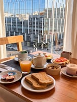 Bandeja de desayuno con tostadas, fruta, té y un pequeño recipiente con frutos secos sobre una mesa junto a una ventana con vistas a la ciudad.
