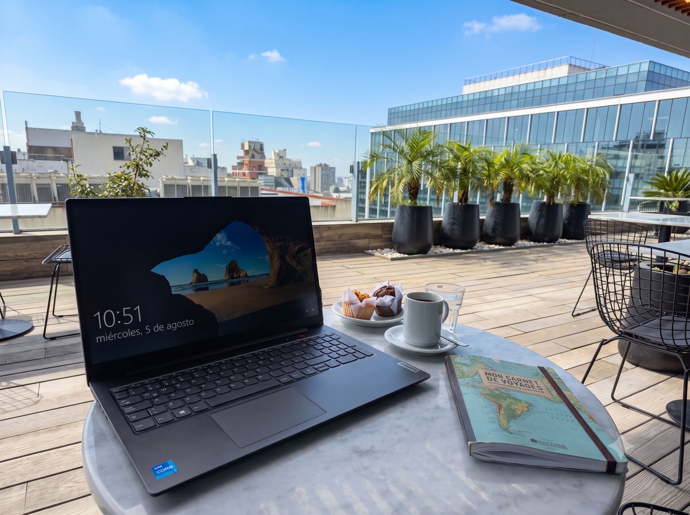 Una laptop sobre una mesa con una taza de café y pasteles, un libro, y vista a un paisaje urbano con edificios y palmeras.