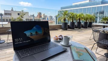 Una laptop sobre una mesa con una taza de café y pasteles, un libro, y vista a un paisaje urbano con edificios y palmeras.