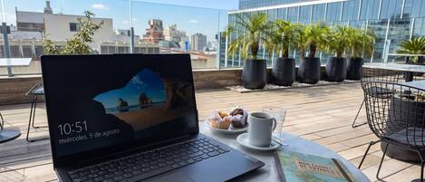 Una laptop sobre una mesa con una taza de café y pasteles, un libro, y vista a un paisaje urbano con edificios y palmeras.