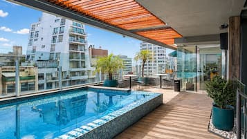 A rooftop infinity pool with a wooden deck, potted plants, and a glass-enclosed dining area.