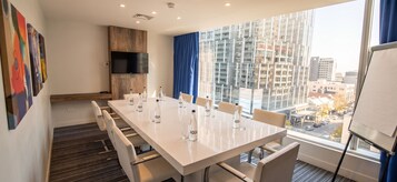 Un salón de conferencias con una larga mesa, sillas, botellas de agua, un televisor y vistas a la ciudad.