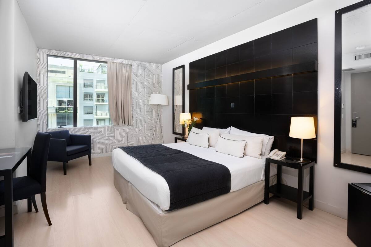 A modern hotel room with a large bed, a desk, a chair, a TV, and a window with a view of buildings.