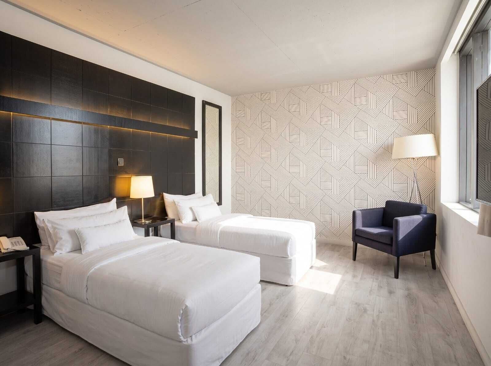 A hotel room with a modern design, featuring a large bed, two bedside tables, a chair, and a patterned wallpaper.