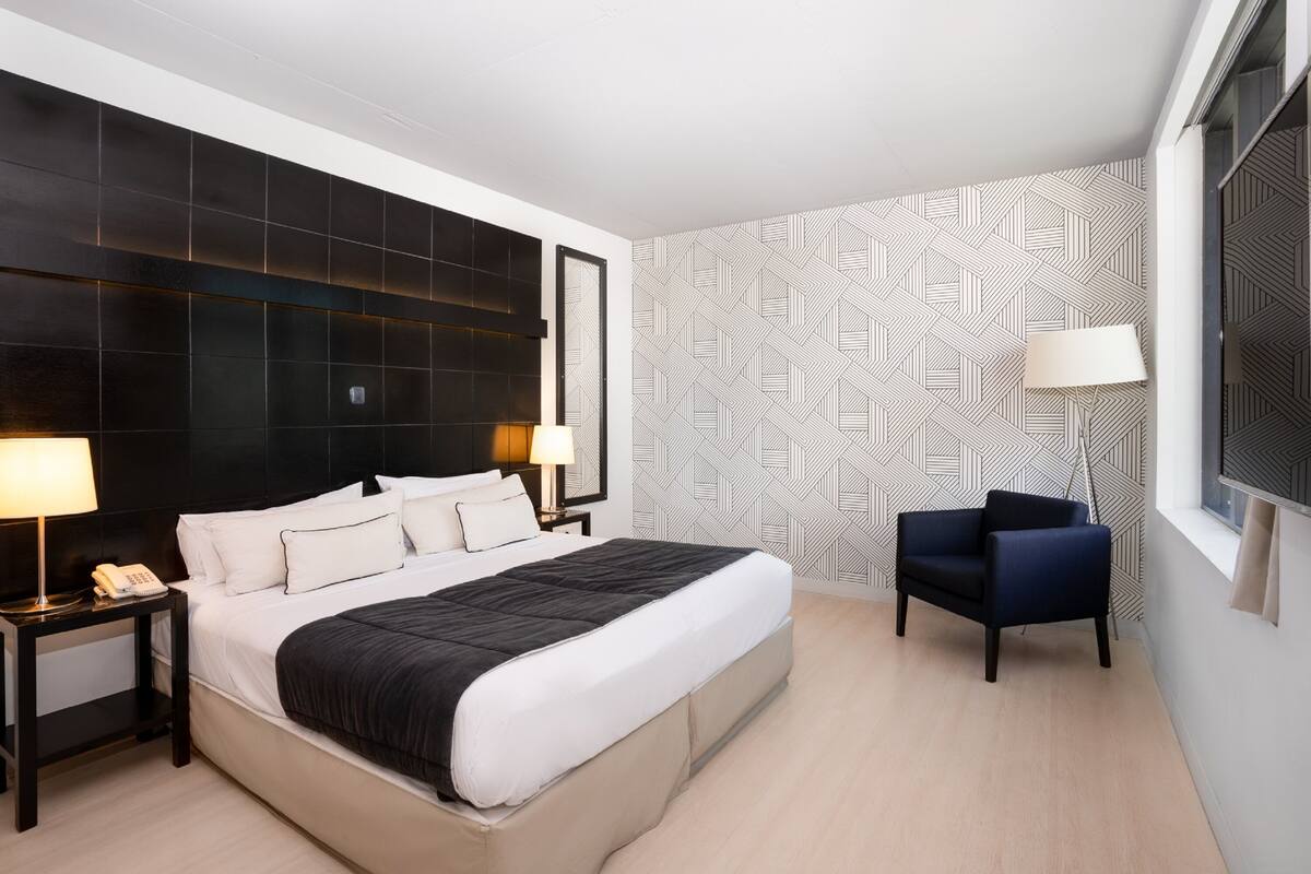 A modern hotel room with a large bed, a nightstand with a phone, a chair, and a patterned wall.