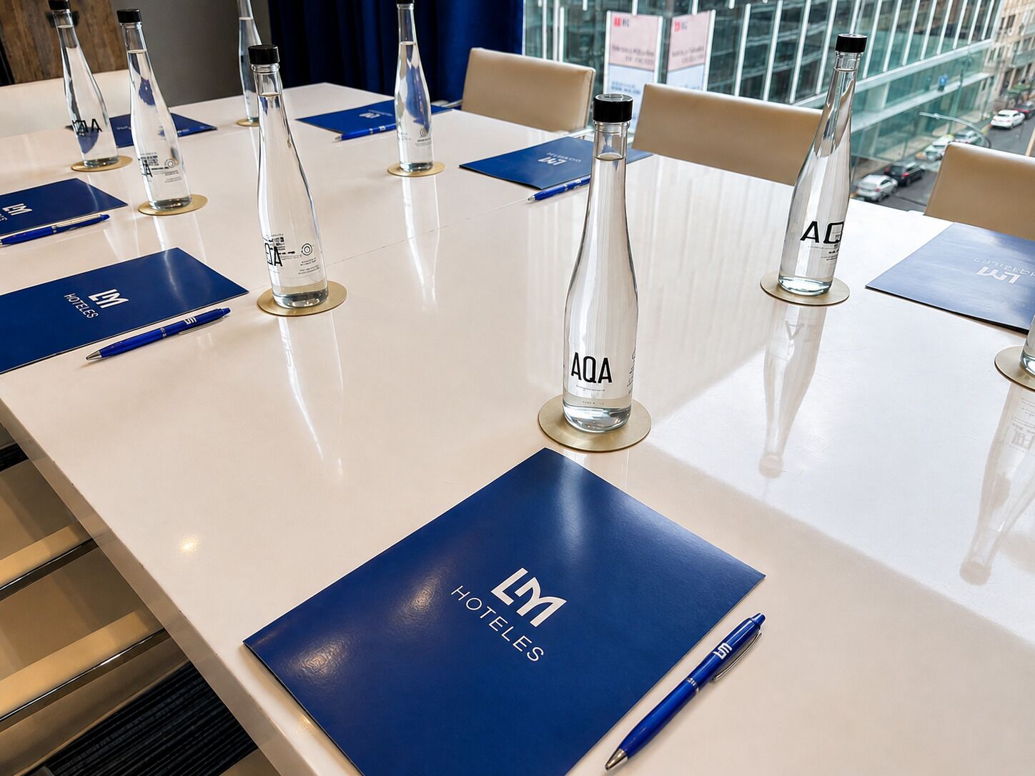 Une table de conférence avec des bouteilles d’eau étiquetées « AQA » et des dossiers bleus portant le logo « LM HOTEL ».