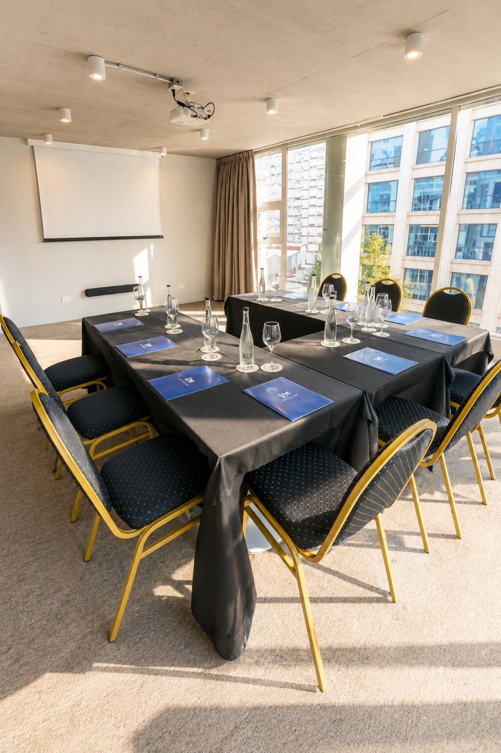 Uma sala de conferências com uma mesa comprida, cadeiras, uma tela de projetor e vista para edifícios através de grandes janelas.