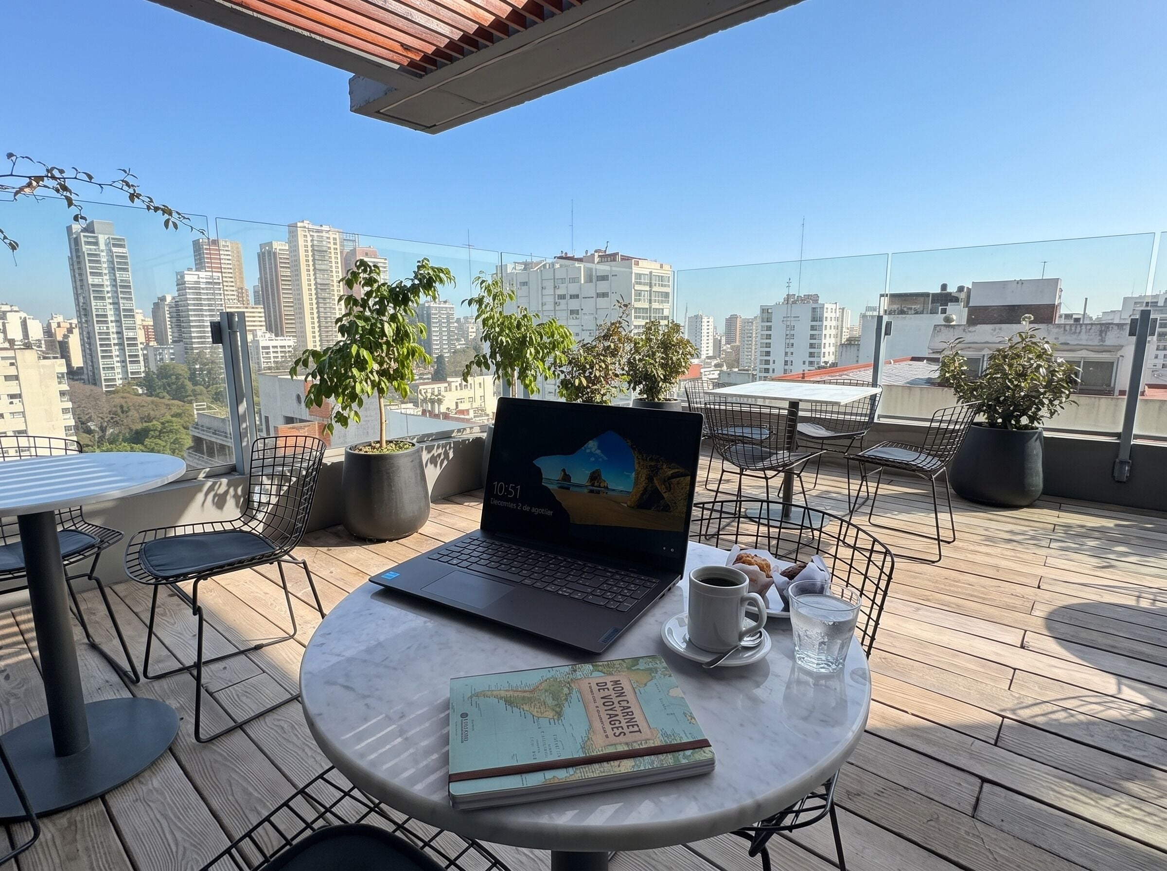 A laptop on a table with a book and a cup, surrounded by chairs and potted plants, with a cityscape view.