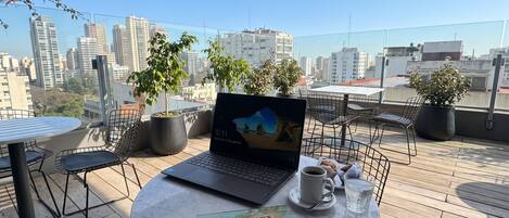 A laptop on a table with a book and a cup, surrounded by chairs and potted plants, with a cityscape view.