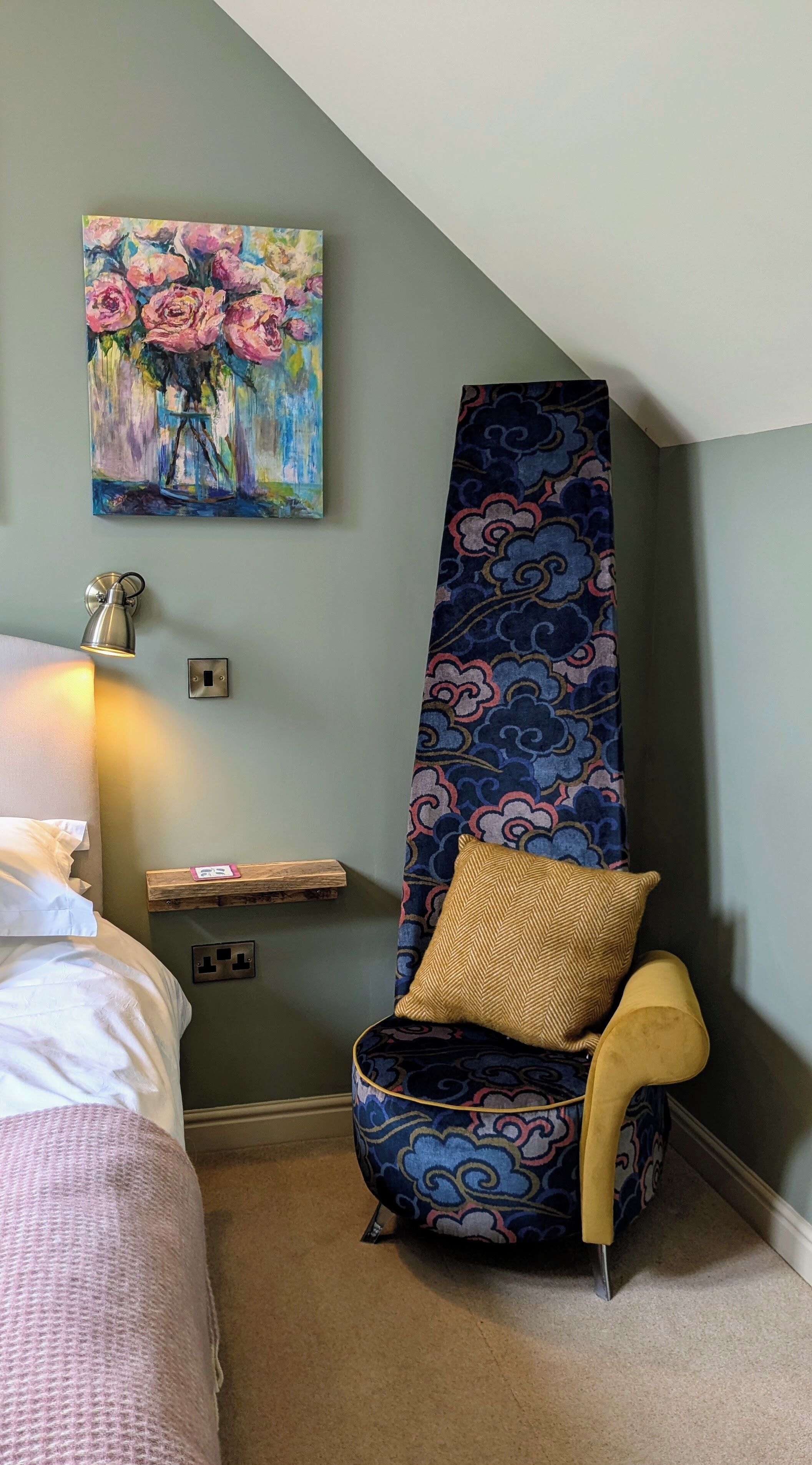 A bedroom with a bed, a chair with a patterned cushion, a painting of flowers, and a wall-mounted lamp.