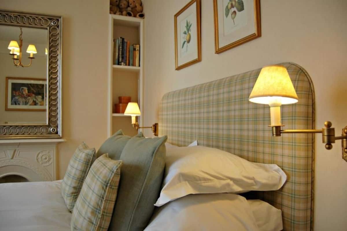 A hotel room with a bed, a headboard lamp, a mirror, and a bookshelf with books and a framed picture.