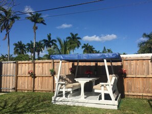 A covered patio area with a swing set and a table, surrounded by a wooden fence and palm trees.