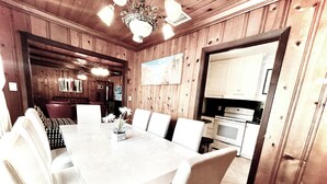 A dining area with a long table and white chairs, wooden paneling on the walls, and a kitchen in the background. - 4BR, 4BA House Beach, Entertainment, Nightlife (Hollywood)