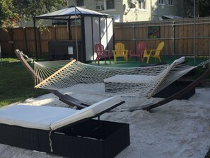 A hammock set up on a sandy surface with a black wicker lounge chair nearby.