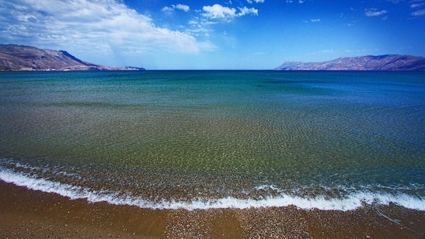 Una playa con agua cristalina, orilla arenosa y montañas distantes bajo un cielo azul.