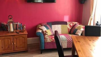 A room with a red wall, a flat-screen TV, a wooden dining table, a sofa, and a bookshelf.