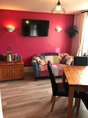 A room with a red wall, a flat-screen TV, a wooden dining table, a sofa, and a bookshelf. - High Ways House (Ilfracombe)