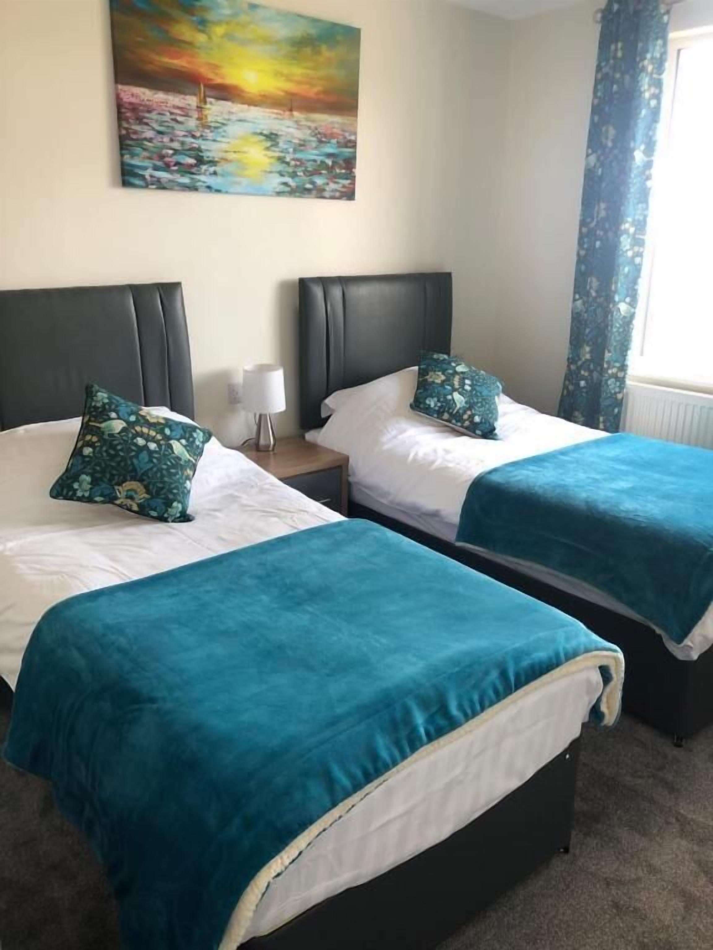 A hotel room with two beds, each with a teal bedspread and matching pillows, a nightstand with a lamp, and a painting of a sunset over water on the wall.