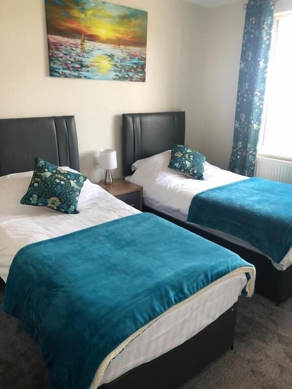 A hotel room with two beds, each with a teal bedspread and matching pillows, a nightstand with a lamp, and a painting of a sunset over water on the wall. - High Ways House (Ilfracombe)