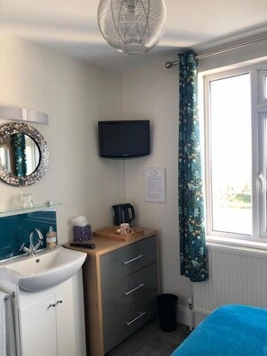 A room with a bed, a TV mounted on the wall, a sink, a mirror, and a window with curtains.