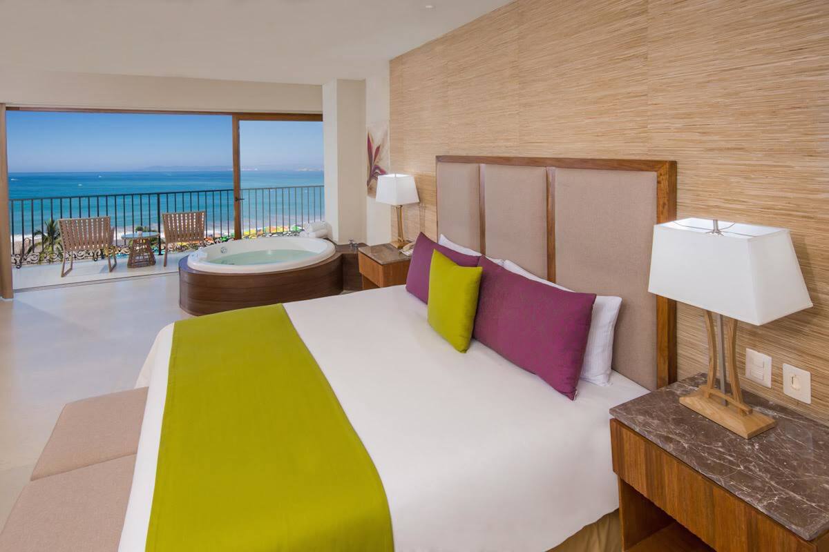 Habitación de hotel con una cama grande, vista al océano, balcón con sillas y un jacuzzi.