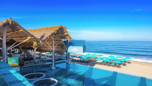 Piscina infinita con vistas a la playa, estructuras de techo de paja y sillones de descanso con sombrillas.