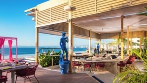 Un área de comedor al aire libre con vistas al mar, que incluye una estatua, mesas, sillas y sombrillas.