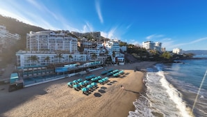 Un hotel frente a la playa con varios pisos, una zona de piscina y sombrillas.