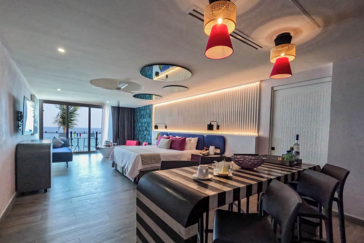 Habitación de hotel moderna con una cama grande, una mesa de comedor y un balcón con vista al mar.