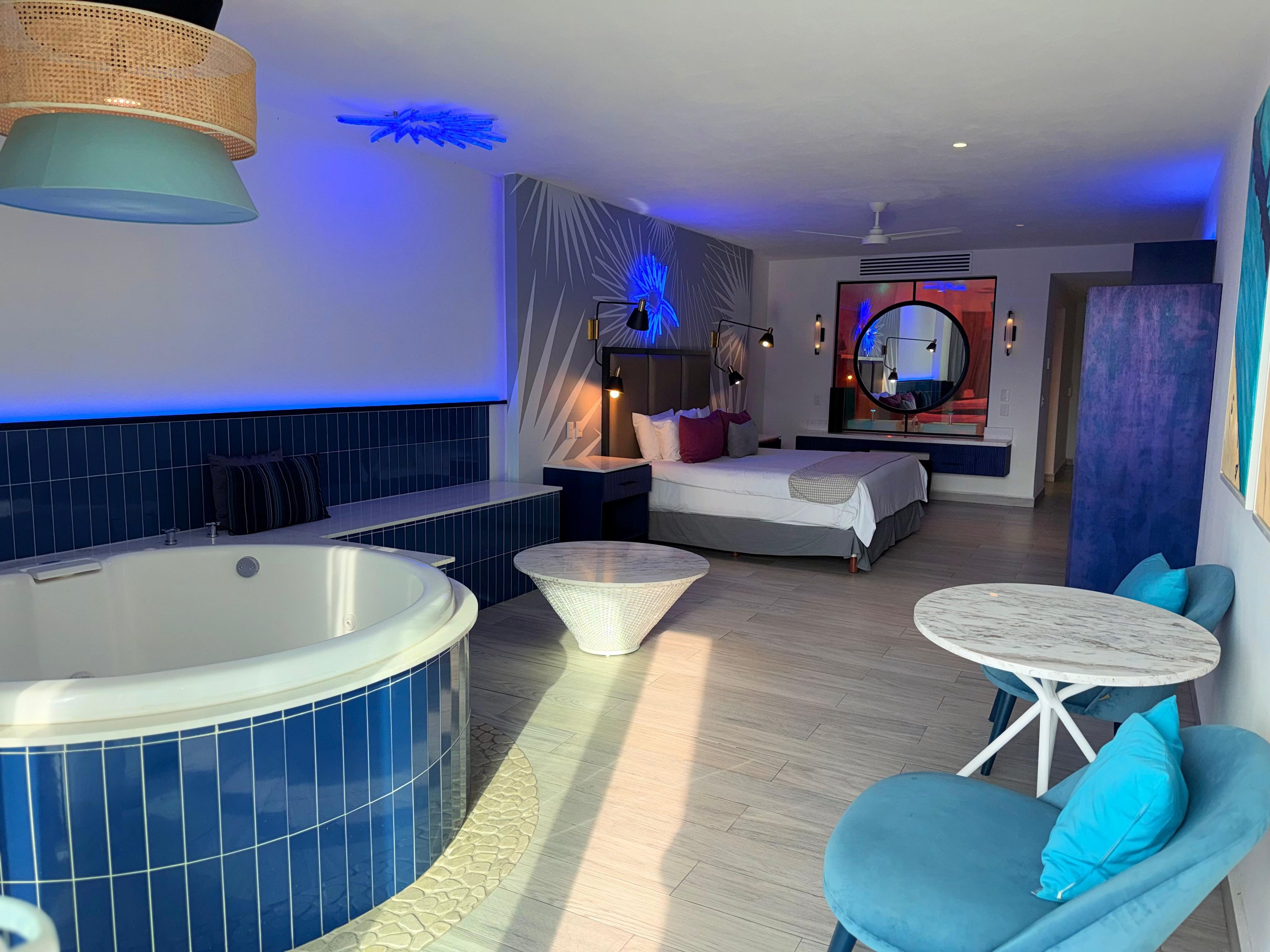 A modern hotel room with a large bathtub, a bed with a view of the room, a small round table, and a seating area with blue chairs.