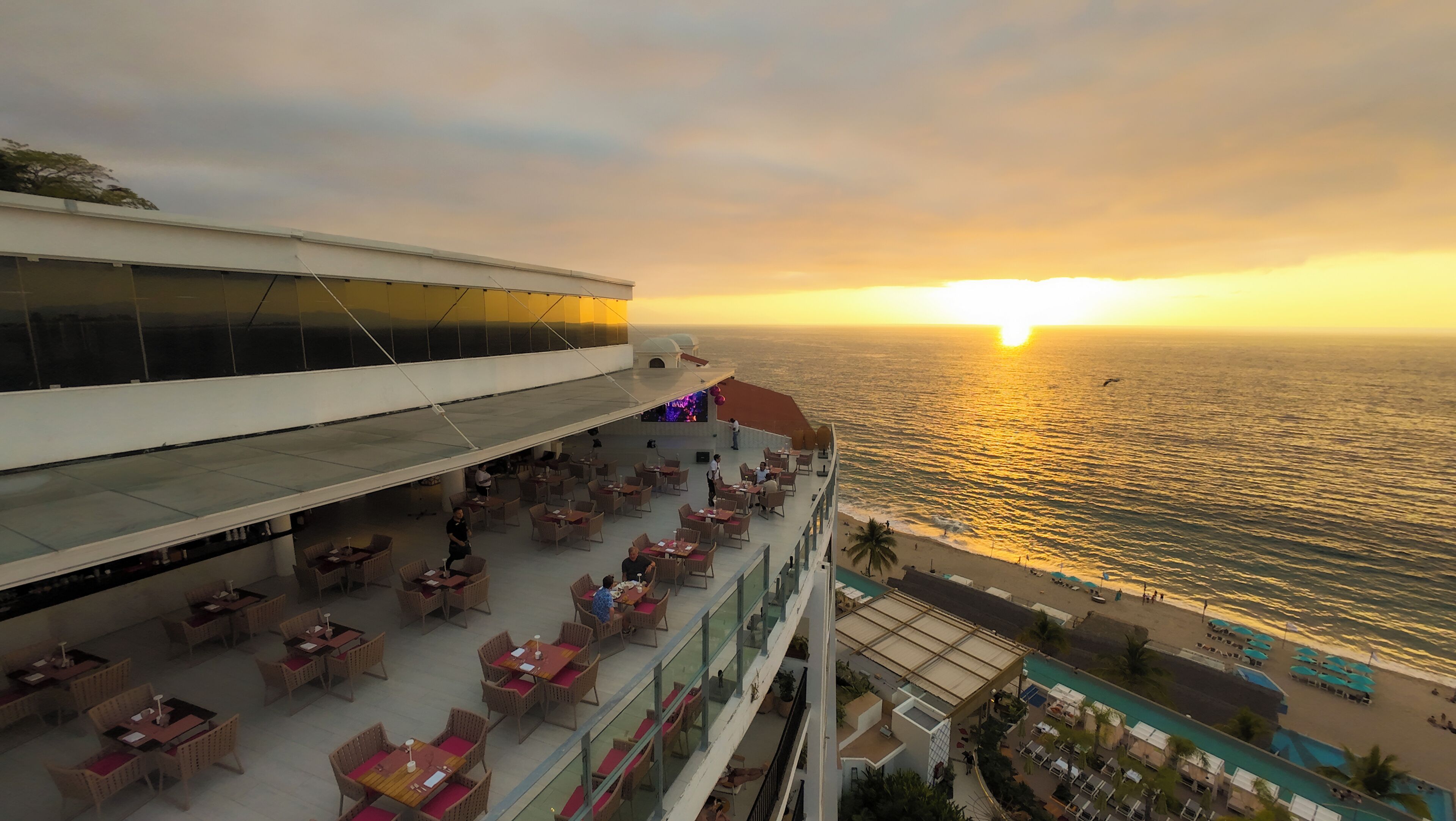 A multi-level outdoor dining area with tables and chairs, overlooking a beach and the ocean at sunset.