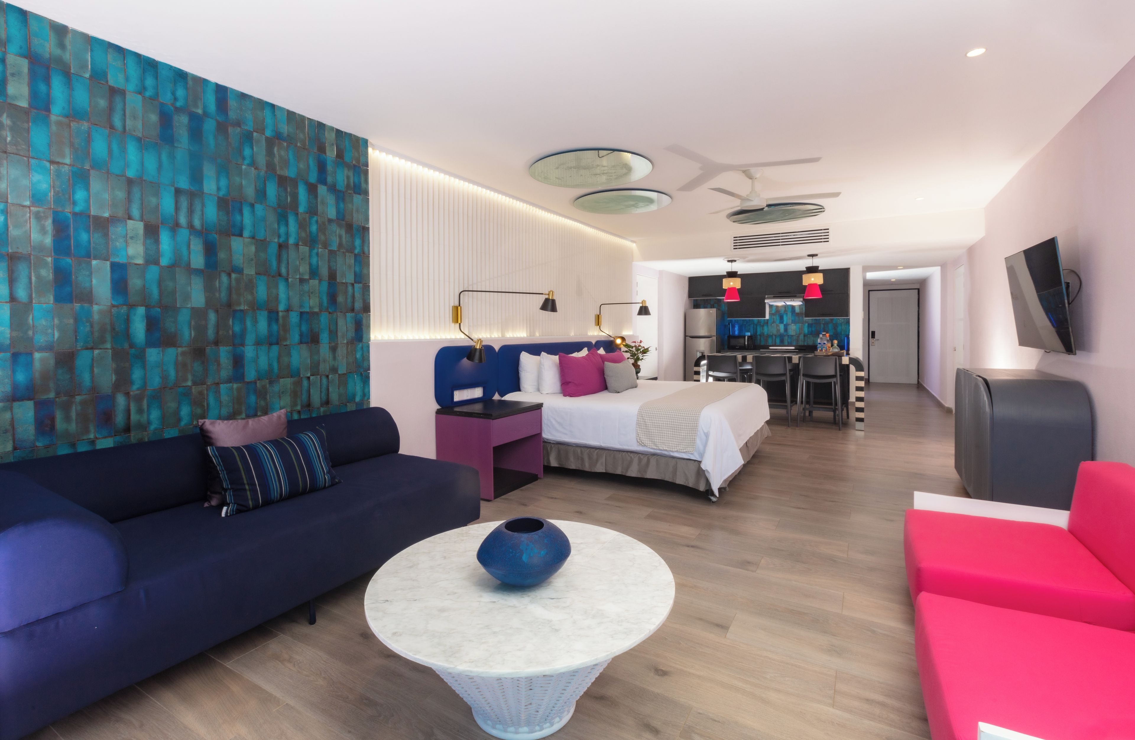 A modern hotel room with a blue sofa, a bed with a white and pink headboard, a small dining area, and a TV mounted on the wall.