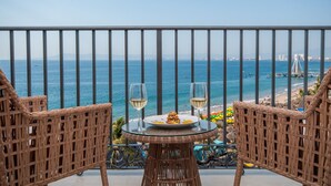 Un balcón con sillas de mimbre, una mesa con un plato de comida y dos copas de vino blanco, con vistas a la playa y al mar.