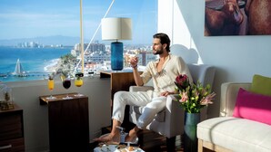 Un hombre sentado junto a una ventana con vistas a un paisaje urbano costero, con una mesa de bebidas y un jarrón de flores.