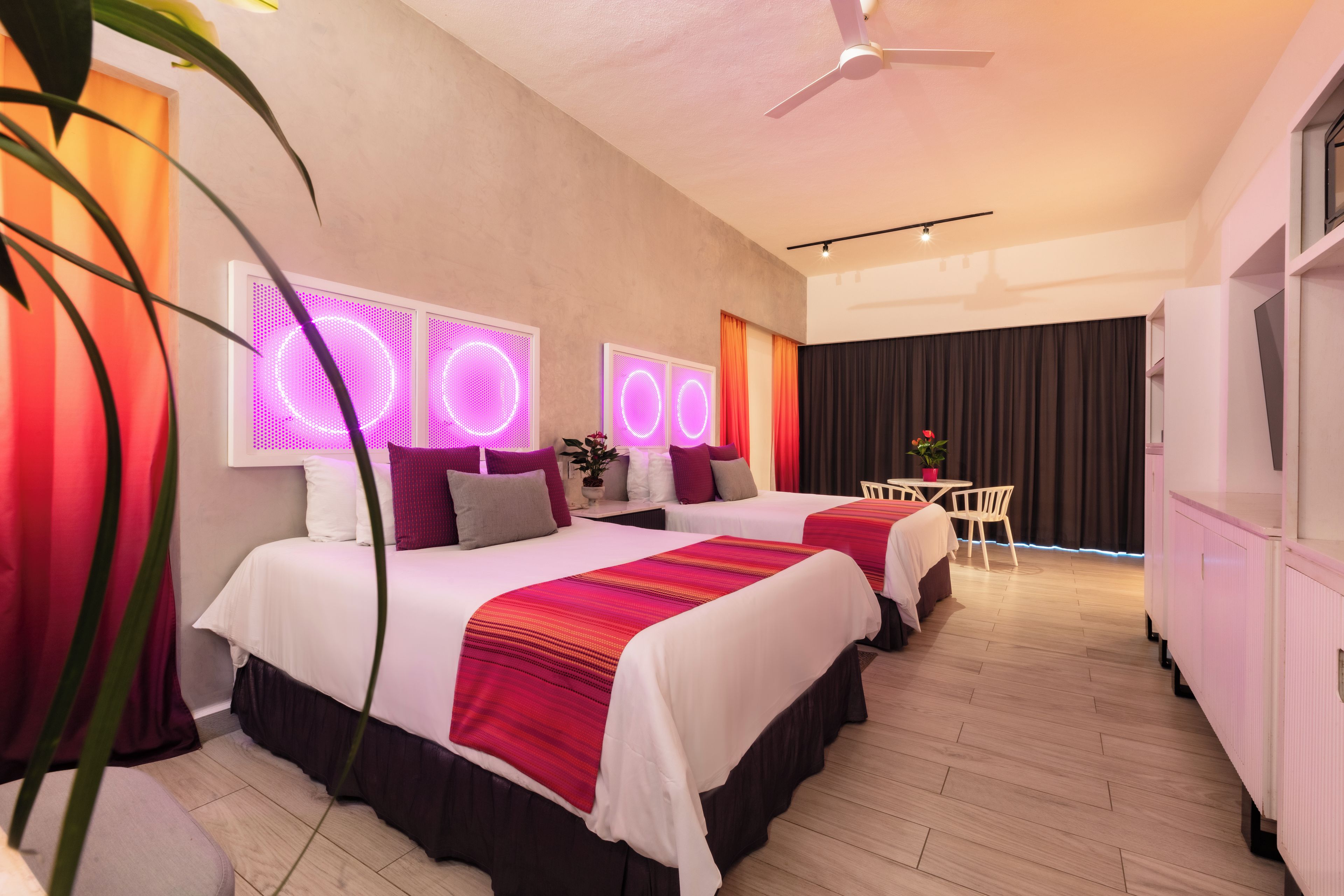 Habitación de hotel con dos camas, un ventilador de techo, una pequeña mesa con sillas y un fondo de cortina oscura.