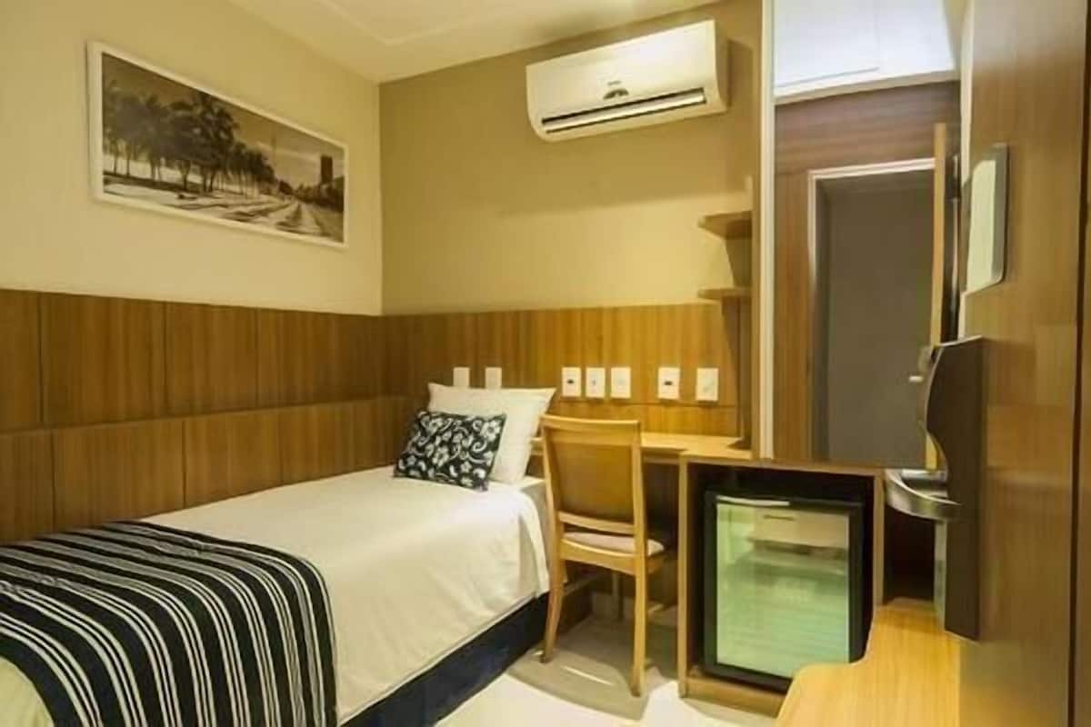 A hotel room with a bed, a desk, a chair, a television, and an air conditioning unit.